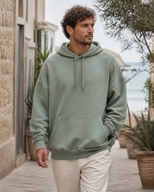 Man wearing an oversized sage organic cotton pullover hoodie in a seaside village